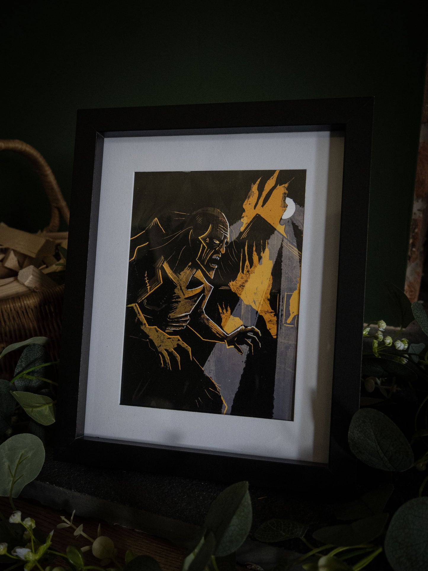 Sophie Atkinson Illustration – The Creature in the Flames Frankenstein print in black frame, full composition view with eucalyptus and wicker basket styling