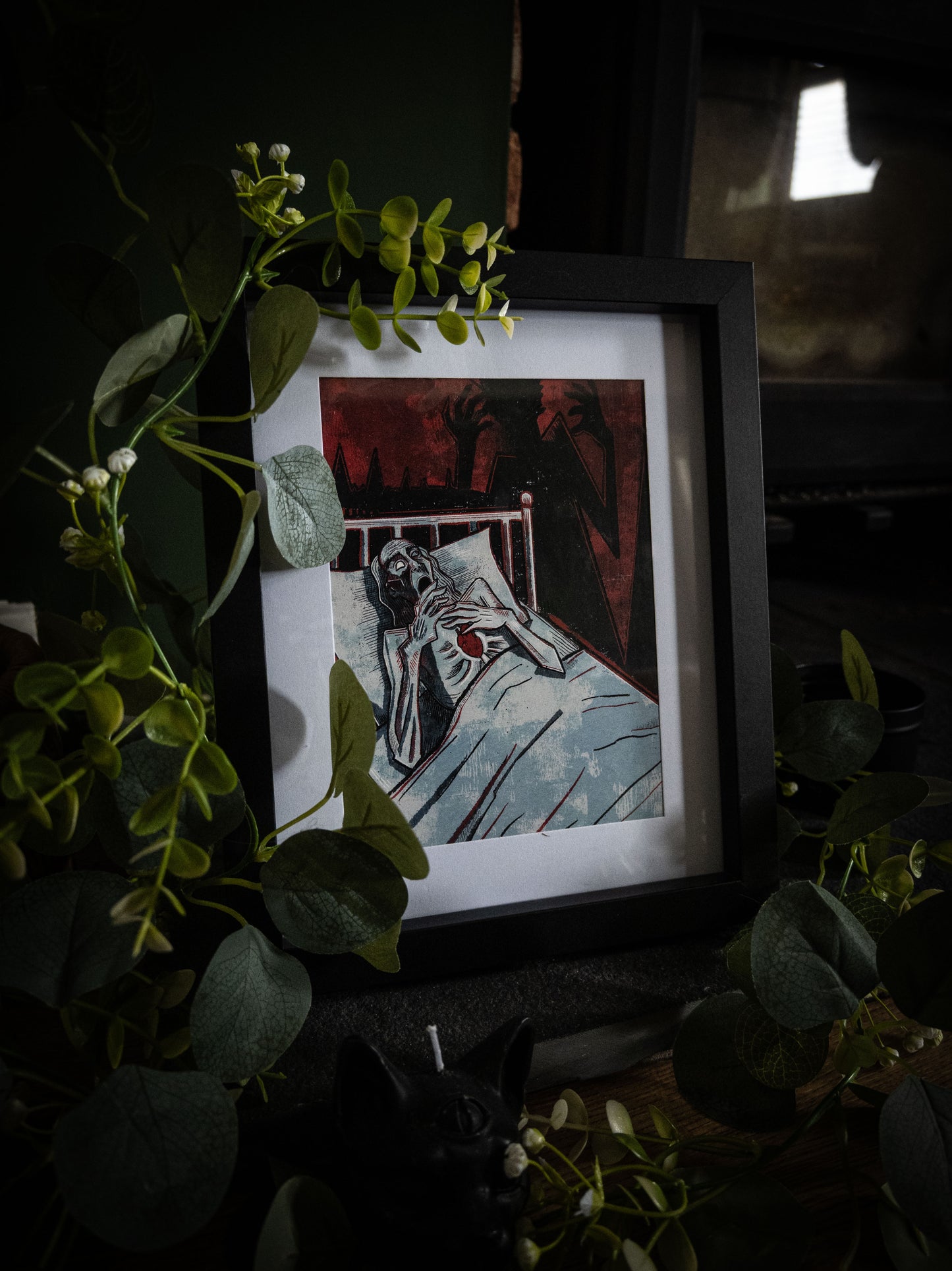 The Tell-Tale Heart Poe anthology art print in black frame, surrounded by lush eucalyptus and white flowers, moody dark atmosphere with crimson and black illustration
