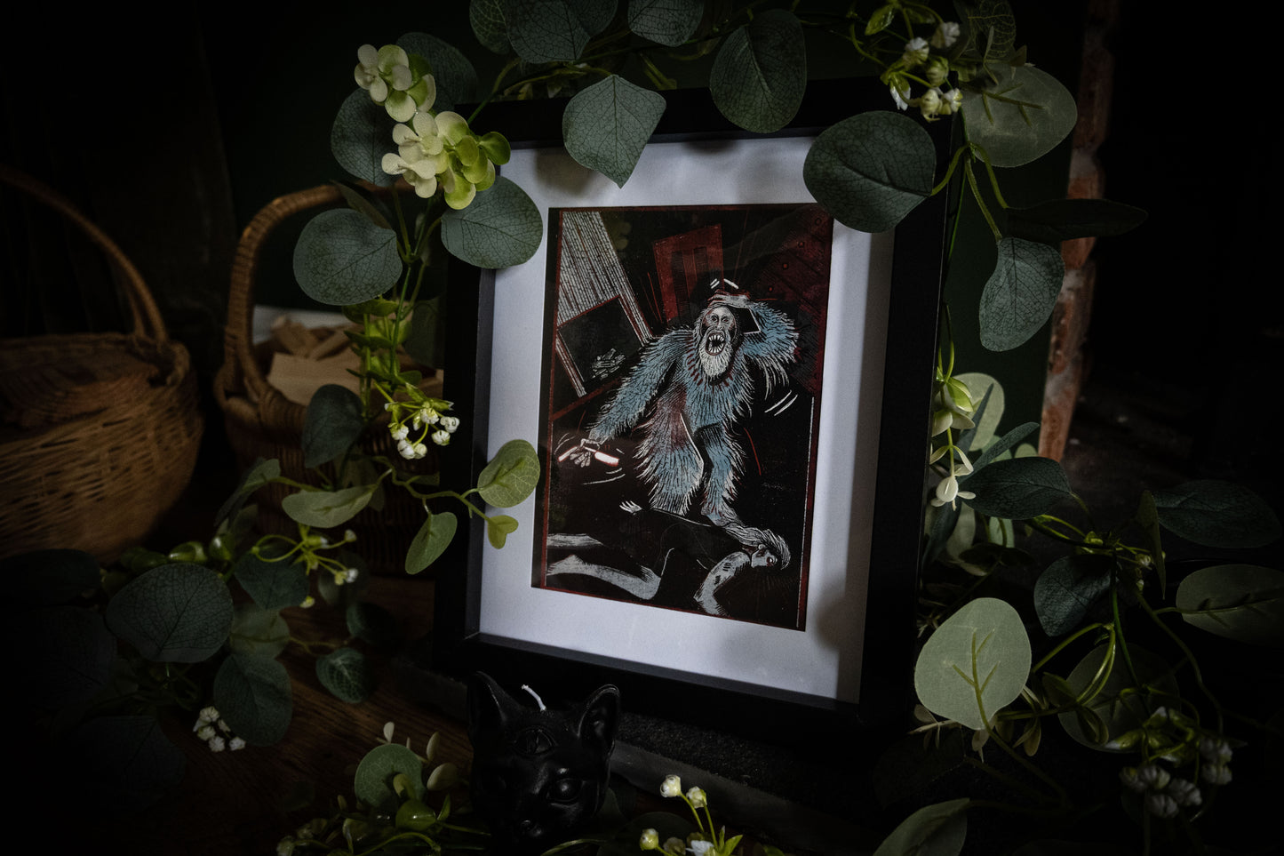Full styled shot of The Murders in the Rue Morgue gothic art print in black frame, surrounded by lush eucalyptus and white flowers with wicker basket, moody dark atmosphere