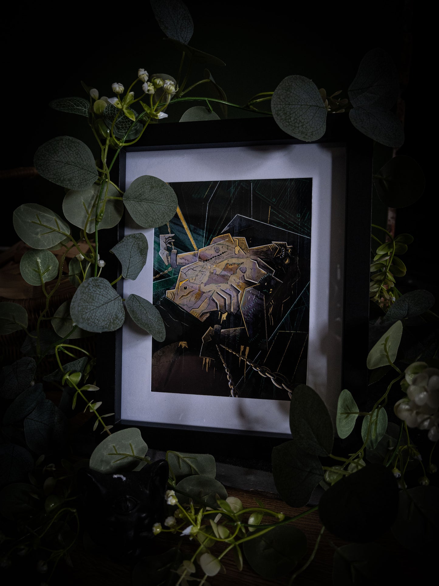 Frankenstein gothic fine art print by Sophie Atkinson in black frame, angled view surrounded by eucalyptus and white flowers on dark wood