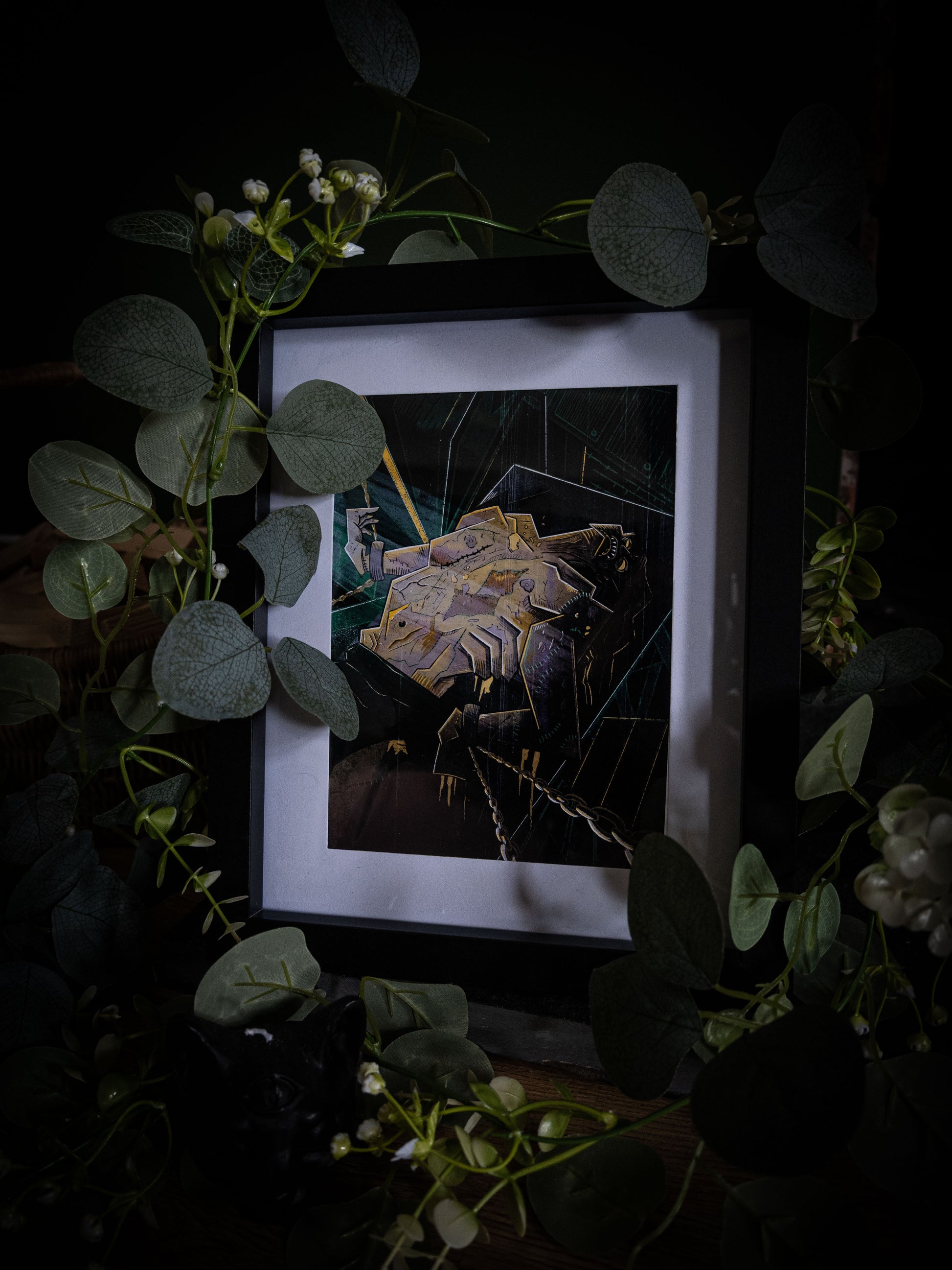 Frankenstein gothic fine art print by Sophie Atkinson in black frame, angled view surrounded by eucalyptus and white flowers on dark wood