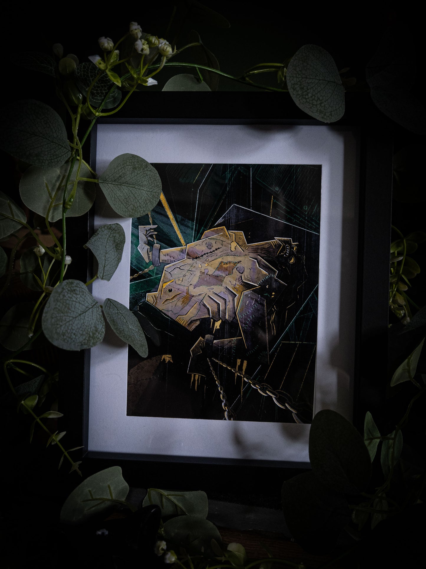 Overhead angled view of The Creature Awakens art print in black frame, dark atmospheric styling with trailing eucalyptus