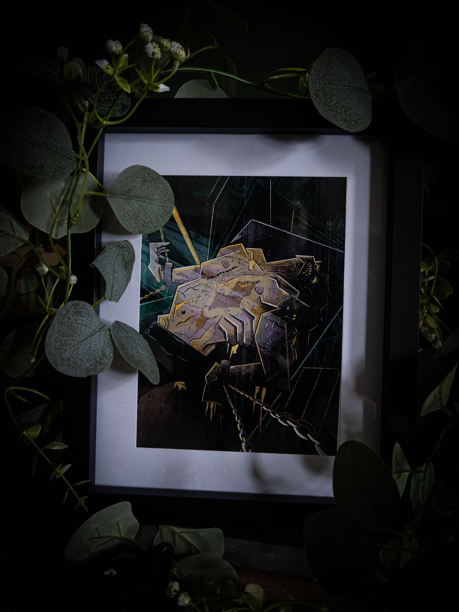 Overhead angled view of The Creature Awakens art print in black frame, dark atmospheric styling with trailing eucalyptus