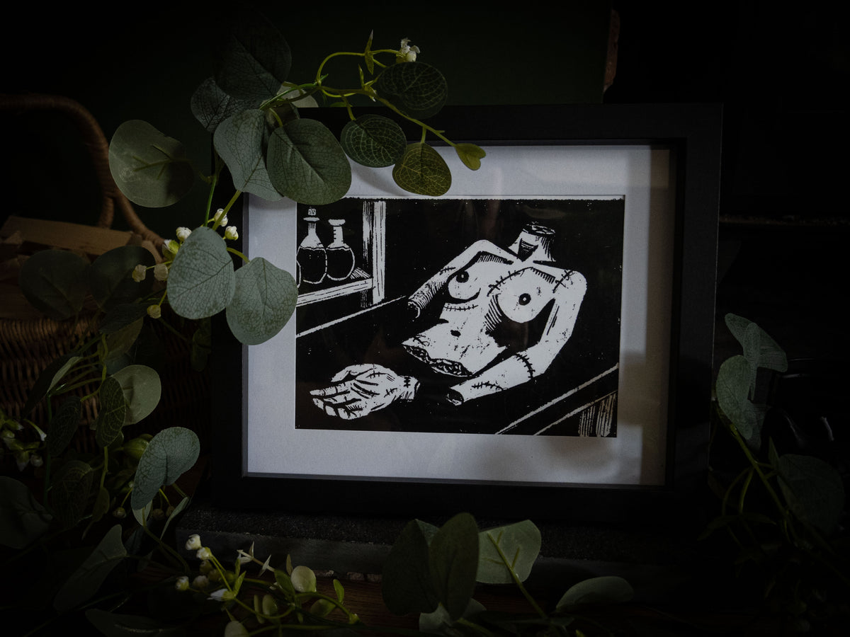 The Workshop Frankenstein art print in black frame surrounded by greenery — gothic horror illustration by Sophie Atkinson