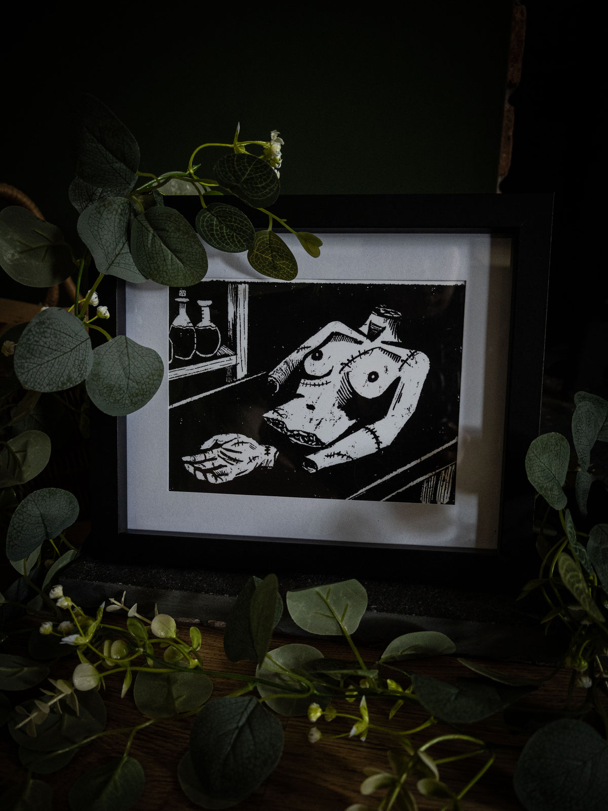 The Workshop giclée print displayed with eucalyptus foliage — Frankenstein creation scene gothic artwork by Sophie Atkinson