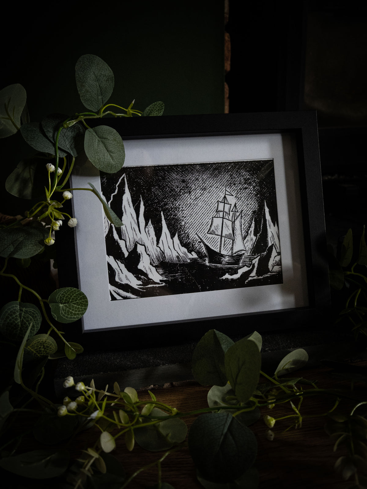 The Arctic Frankenstein art print in black frame surrounded by greenery — ship and icebergs, high-contrast gothic illustration