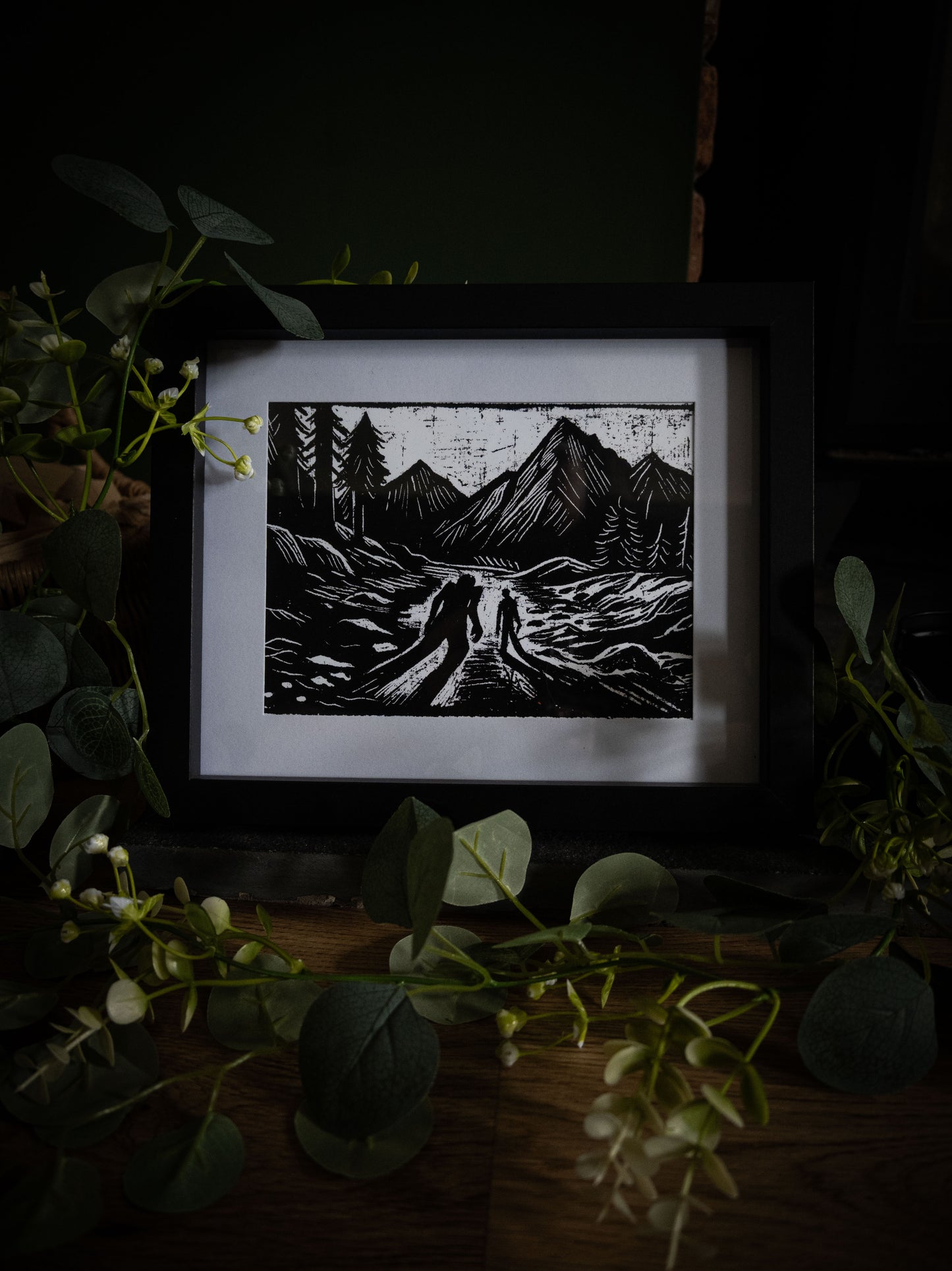 The Alpine Pass Frankenstein art print in black frame surrounded by greenery — two figures among peaks and pines, gothic illustration