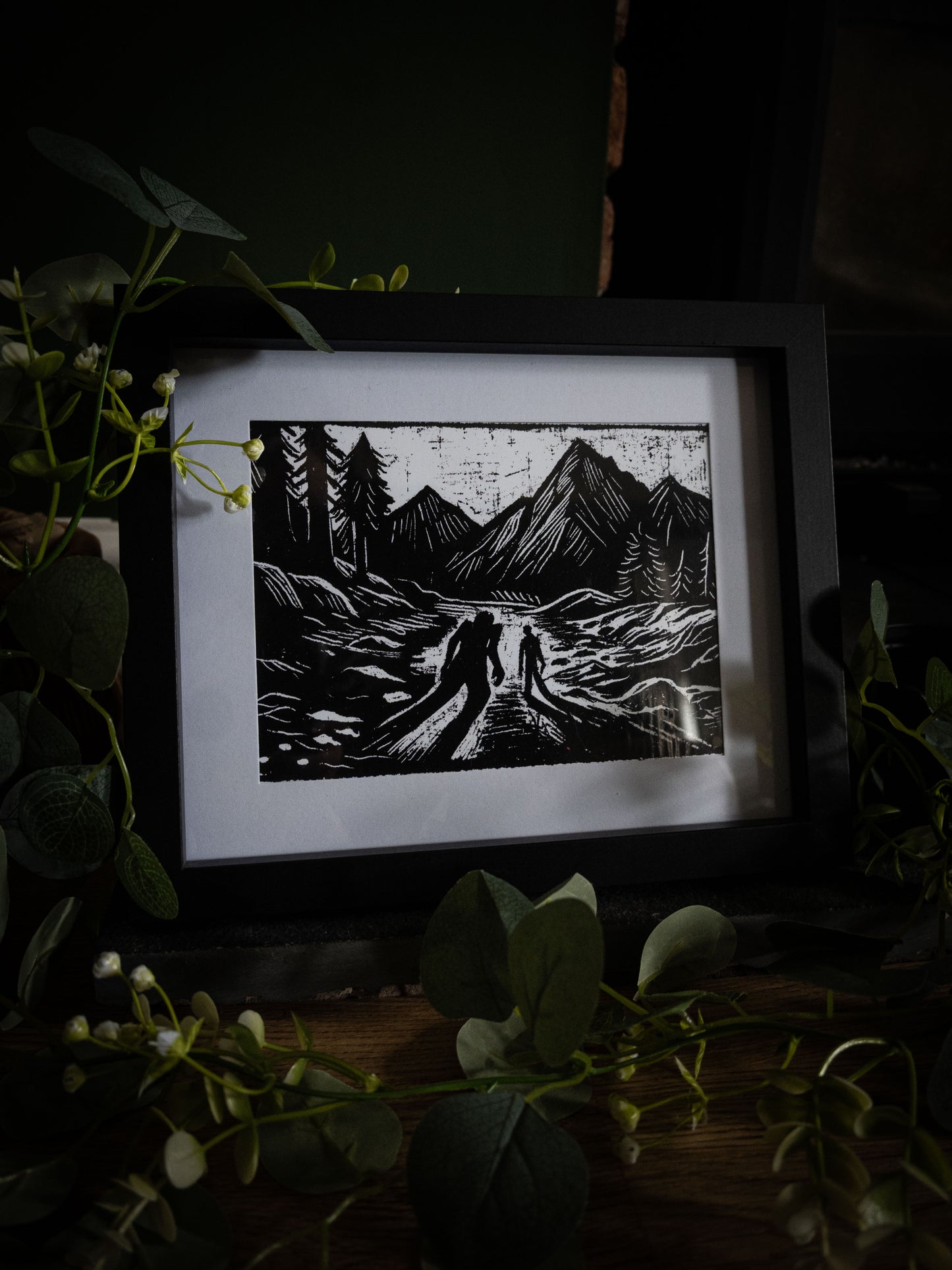 The Alpine Pass art print in black frame with eucalyptus — two silhouettes on a mountain track, bold gothic illustration by Sophie Atkinson
