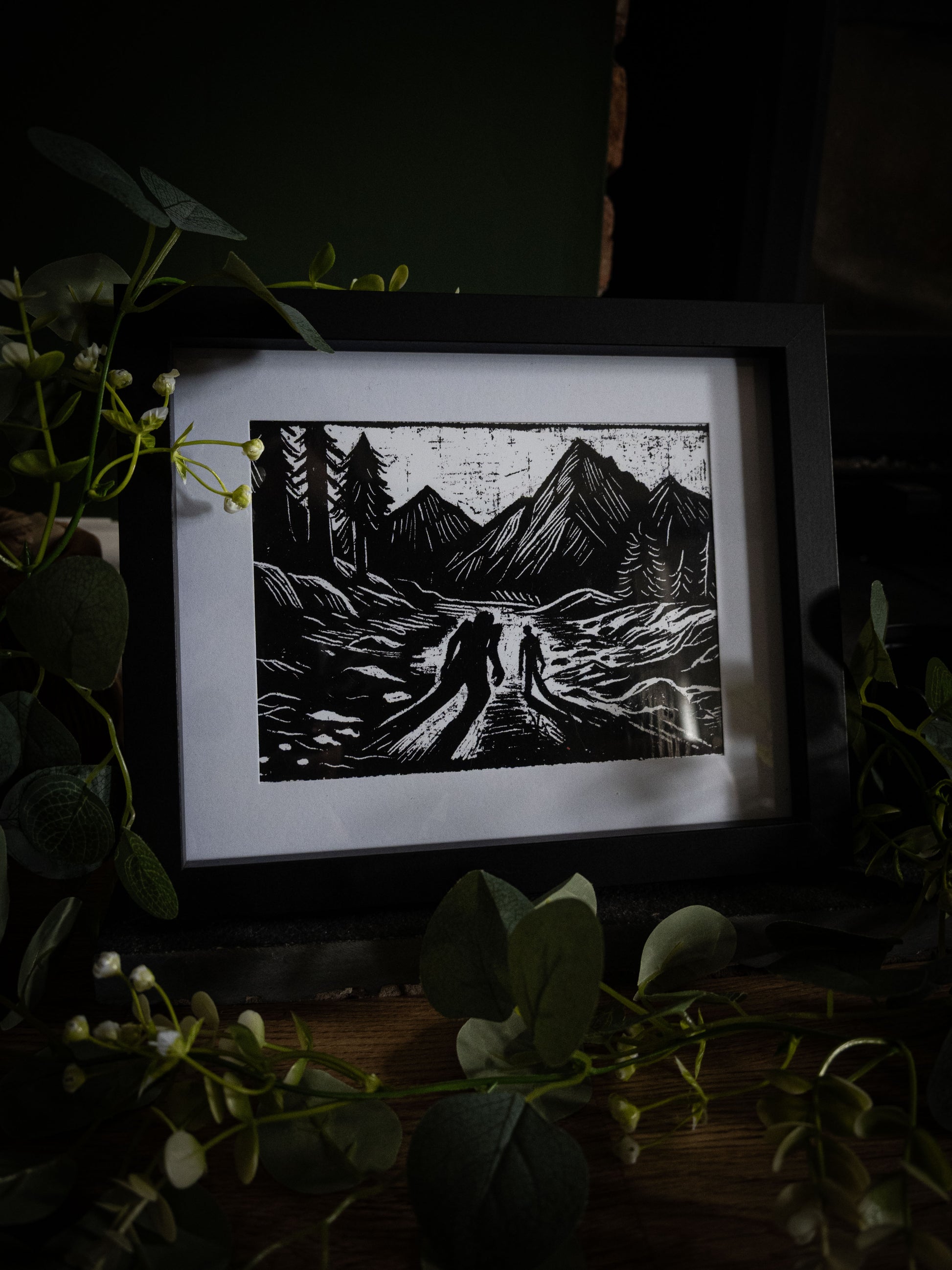The Alpine Pass art print in black frame with eucalyptus — two silhouettes on a mountain track, bold gothic illustration by Sophie Atkinson