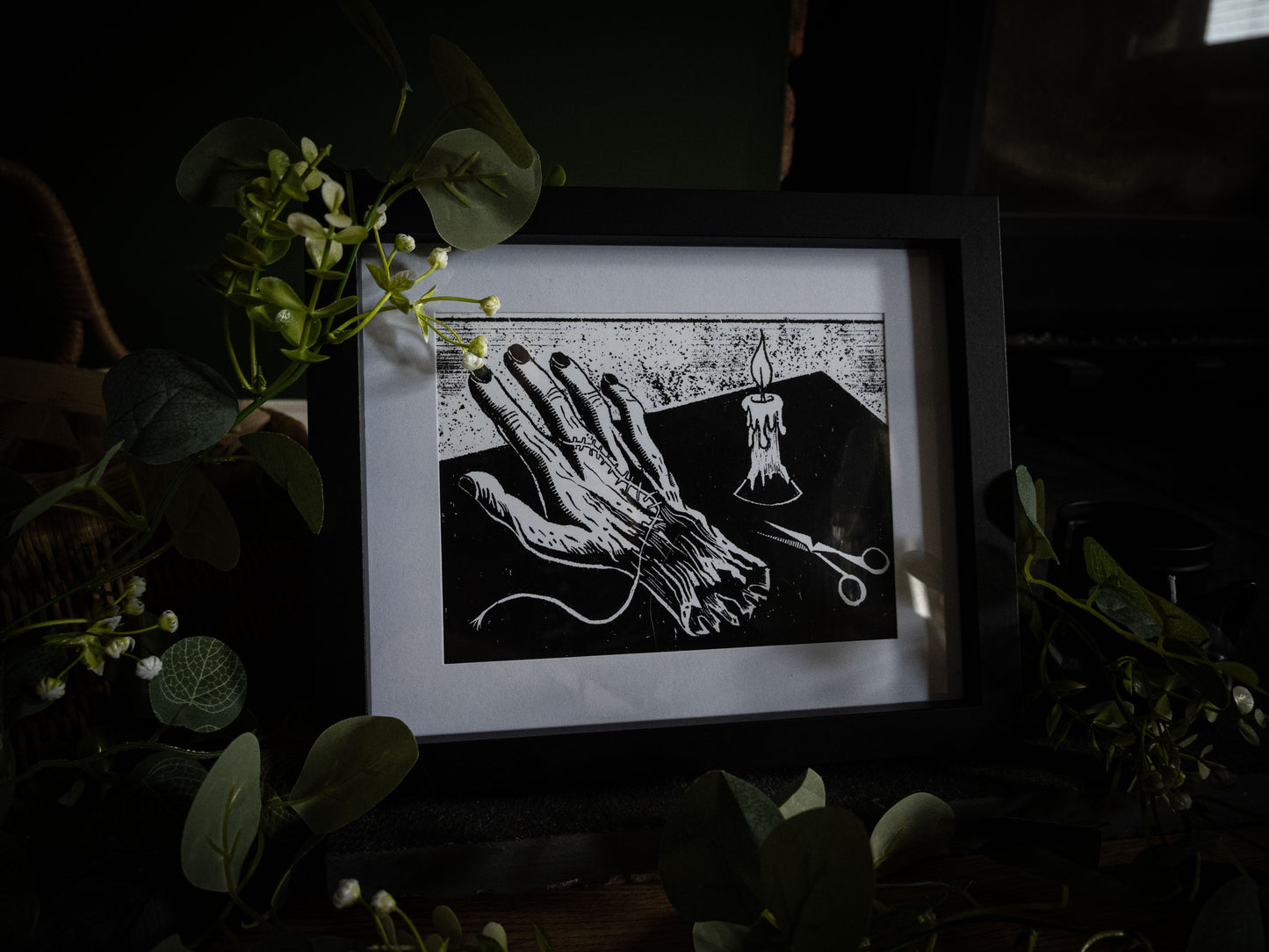 The Stitching giclée print displayed with winter foliage — Frankenstein creation detail, creature's hand with scissors and candle, gothic artwork