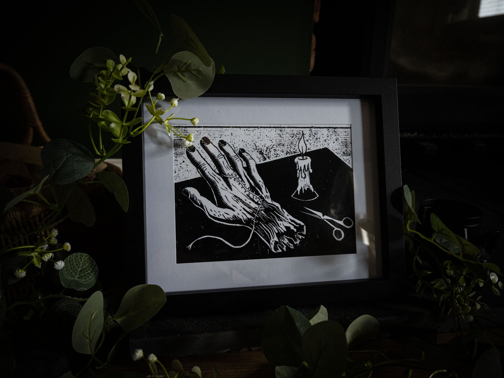 The Stitching giclée print displayed with winter foliage — Frankenstein creation detail, creature's hand with scissors and candle, gothic artwork