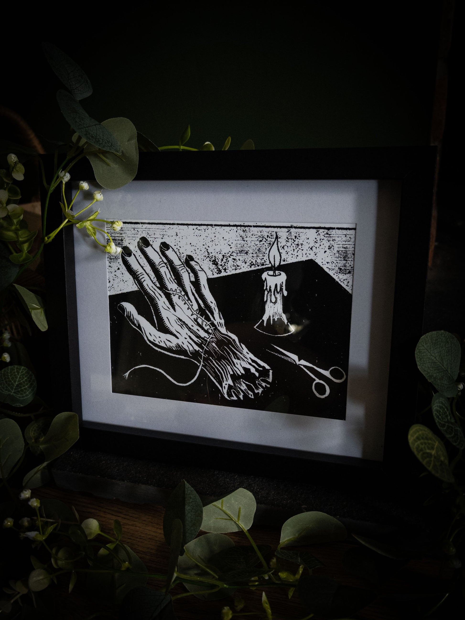 The Stitching art print in black frame with eucalyptus — needle and thread through assembled hand, bold gothic illustration by Sophie Atkinson