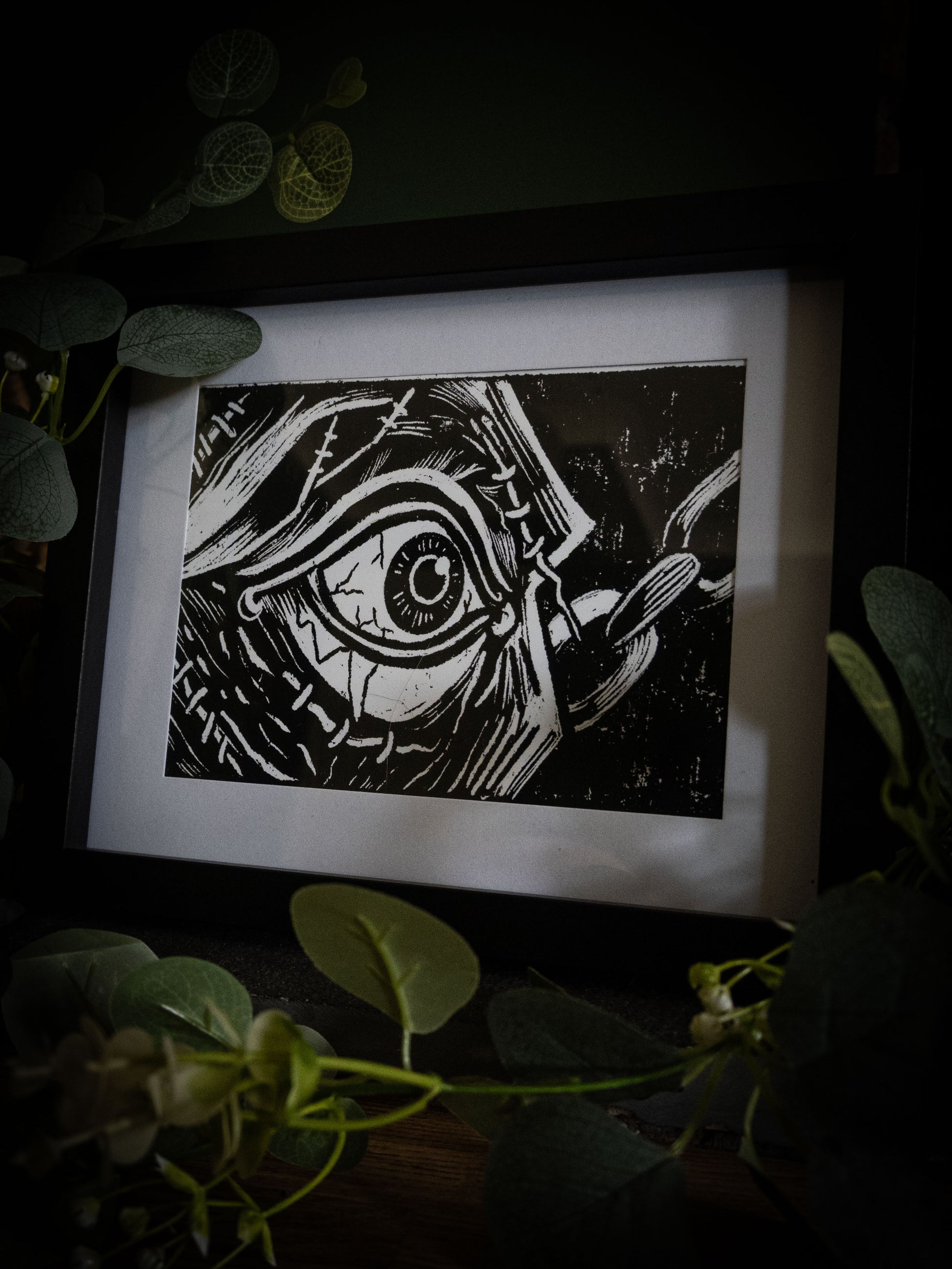 The First Sight art print in black frame with eucalyptus — extreme close-up of the creature's awakening eye, bold gothic illustration by Sophie Atkinson