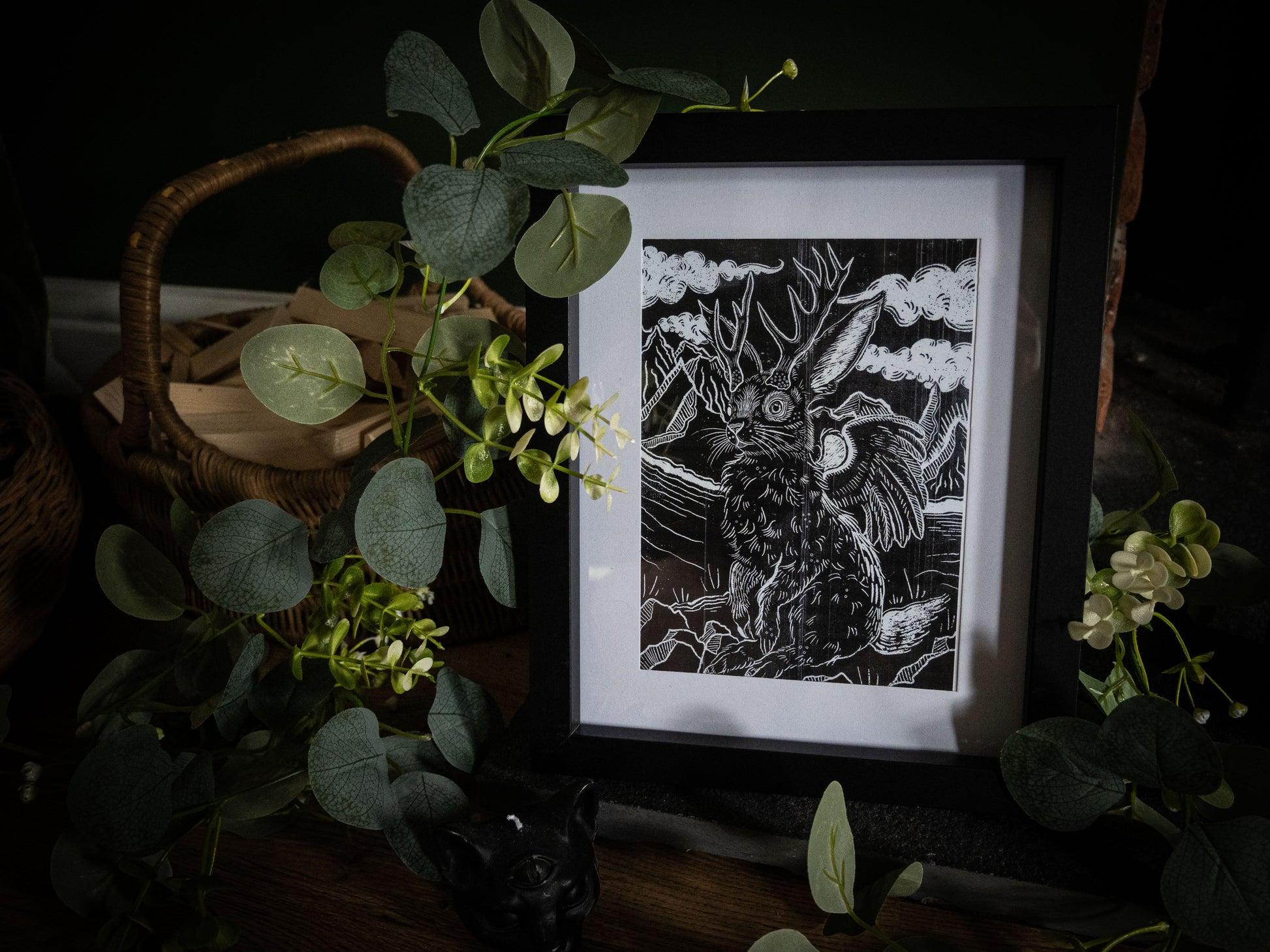 Jackalope gothic fine art print by Sophie Atkinson, framed in black, displayed on a dark wooden surface surrounded by eucalyptus and foliage