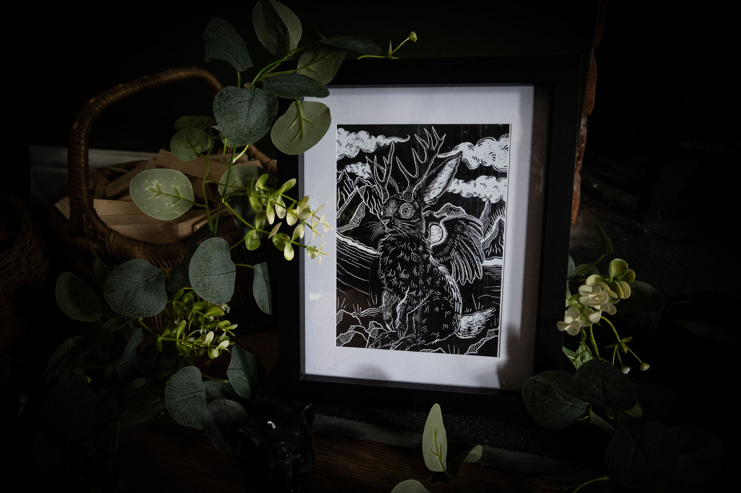 Close-up of Jackalope woodblock-style art print in black frame, antlered hare with wings against a stormy sky, surrounded by green foliage