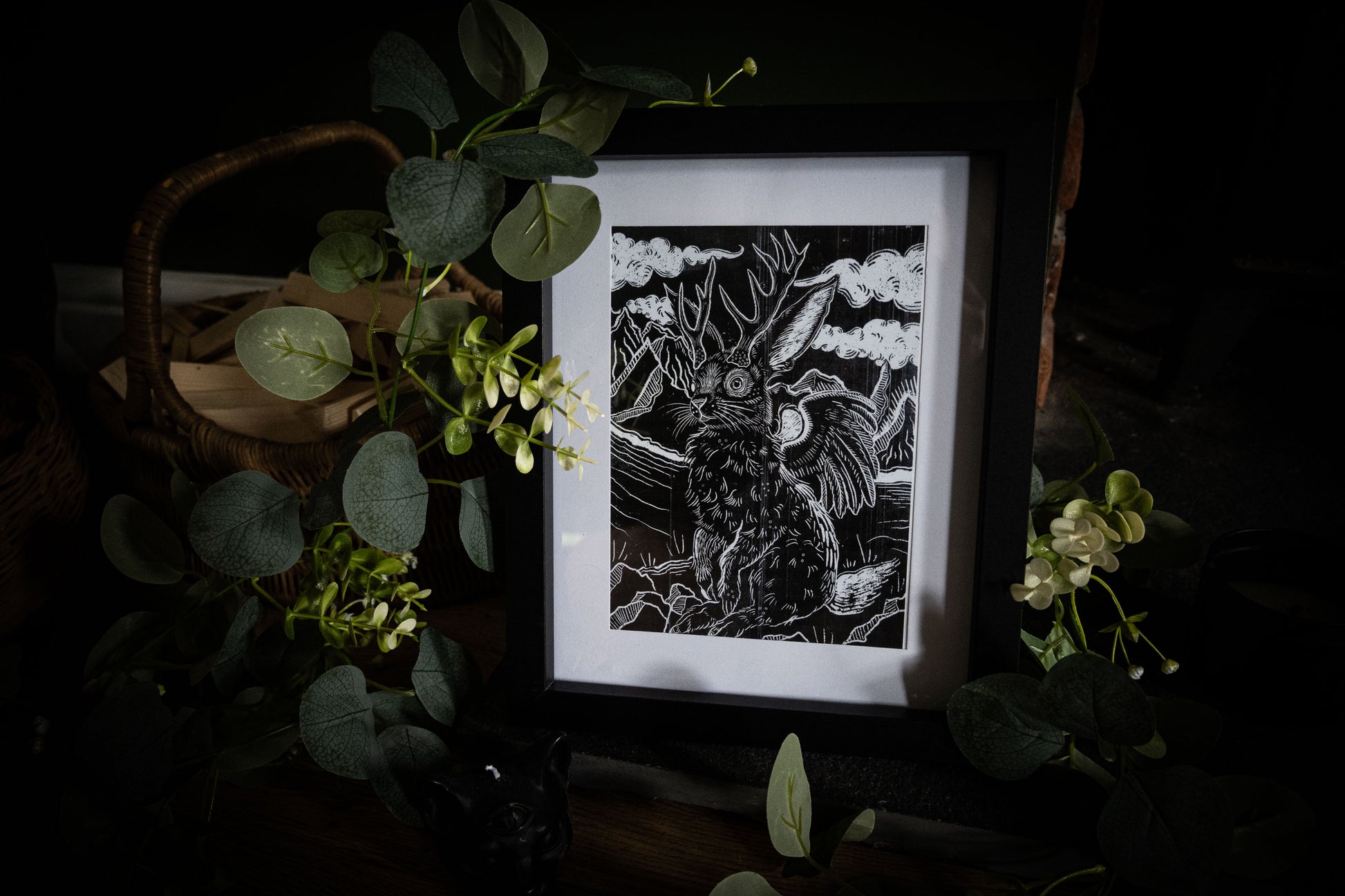 Close-up of Jackalope woodblock-style art print in black frame, antlered hare with wings against a stormy sky, surrounded by green foliage