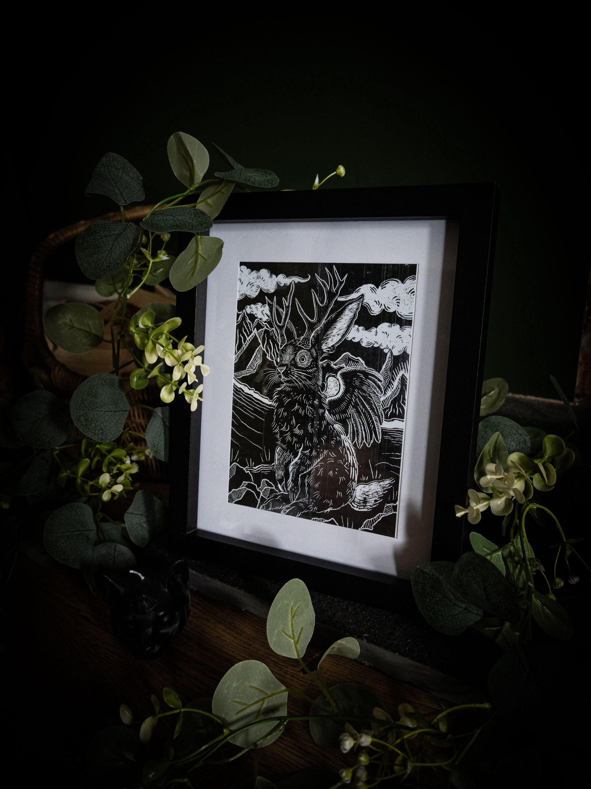 Full view of Jackalope gothic illustration print in black frame, styled with eucalyptus and botanical props on dark wood