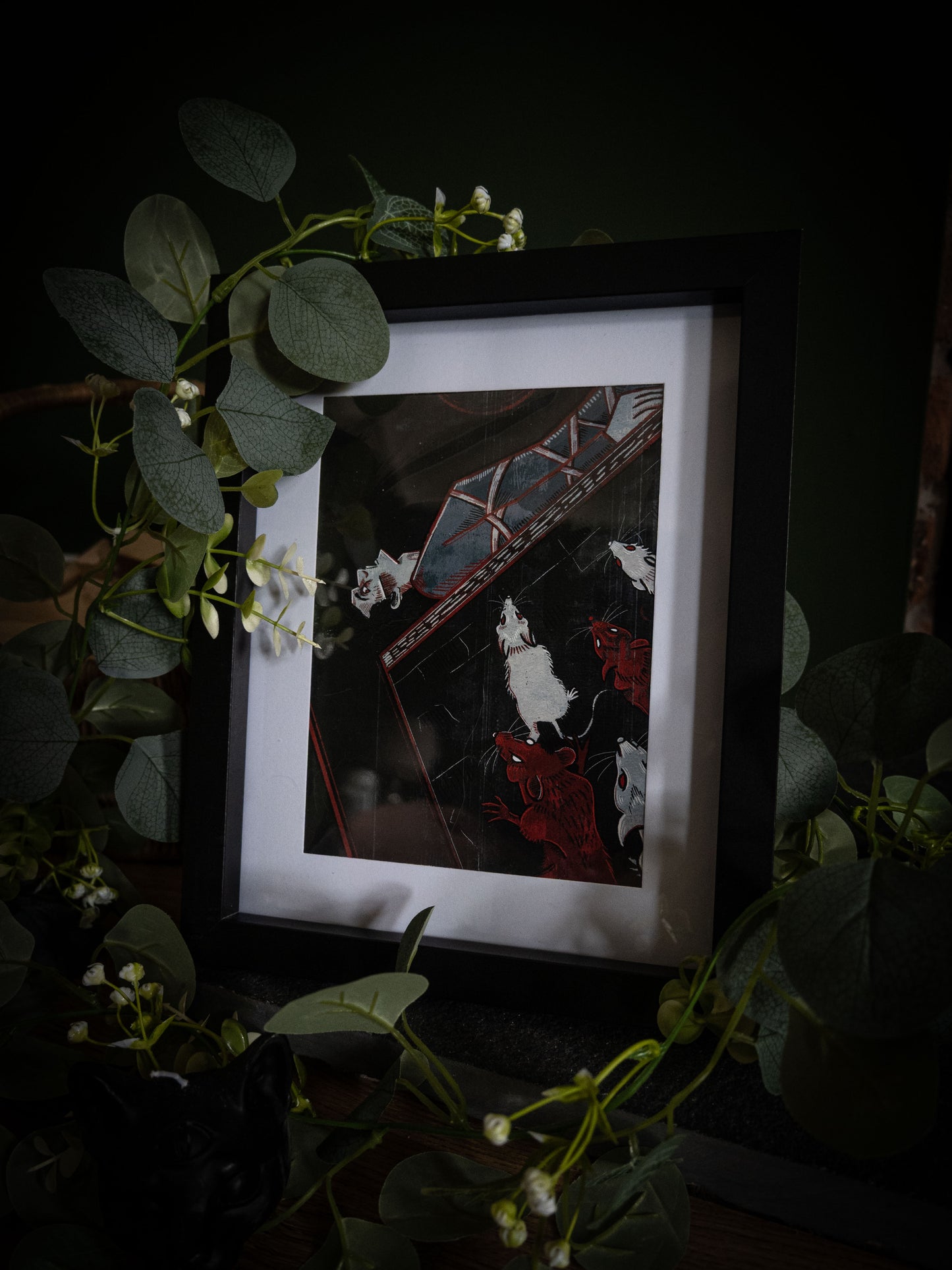The Pit and the Pendulum gothic art print by Sophie Atkinson, framed in black on a dark surface surrounded by eucalyptus, showing pendulum blade and rats in deep black and crimson