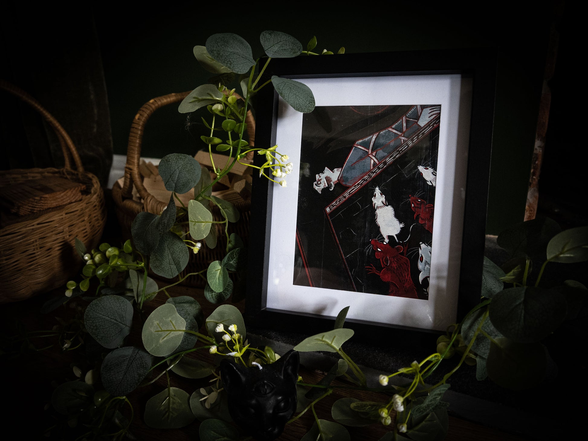The Pit and the Pendulum Edgar Allan Poe art print in black frame, angled view surrounded by eucalyptus and white flowers on dark surface with wicker basket