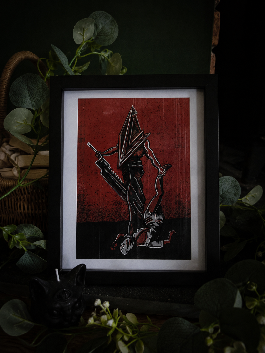 An art print featuring a stylized black and white illustration of pyramid head from silent hill holding a knife against a textured red background, displayed in a dark, moody setting 