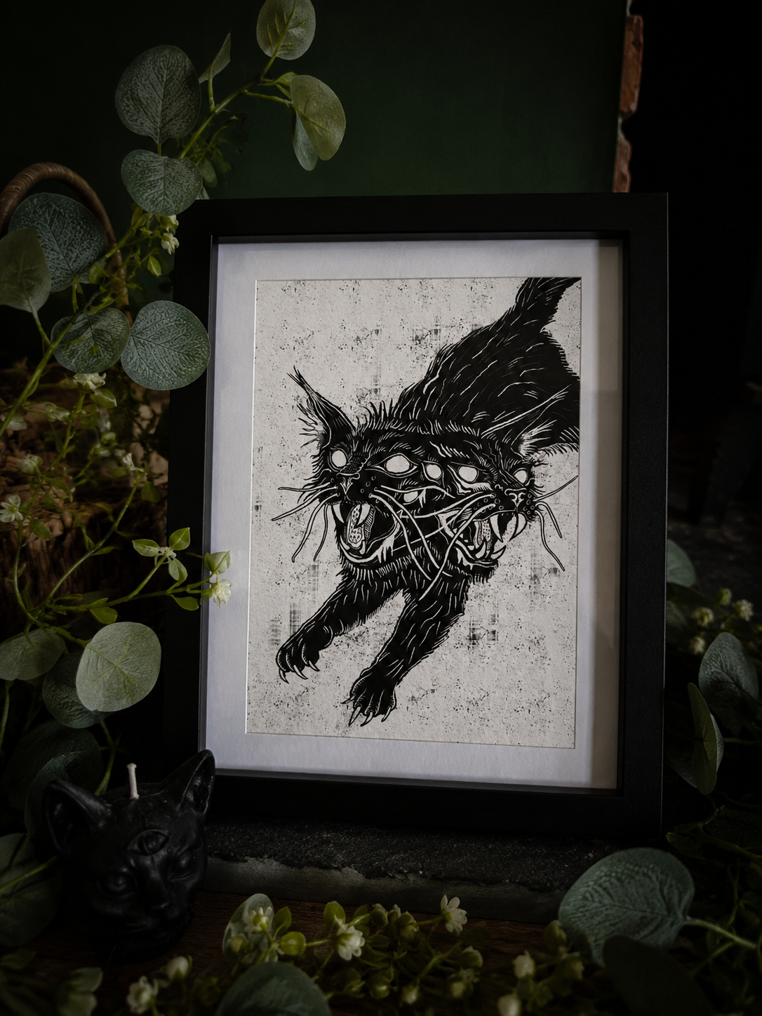 A black and white art print of a snarling two headed cat, styled with green foliage and a small black cat figurine in a dark setting.