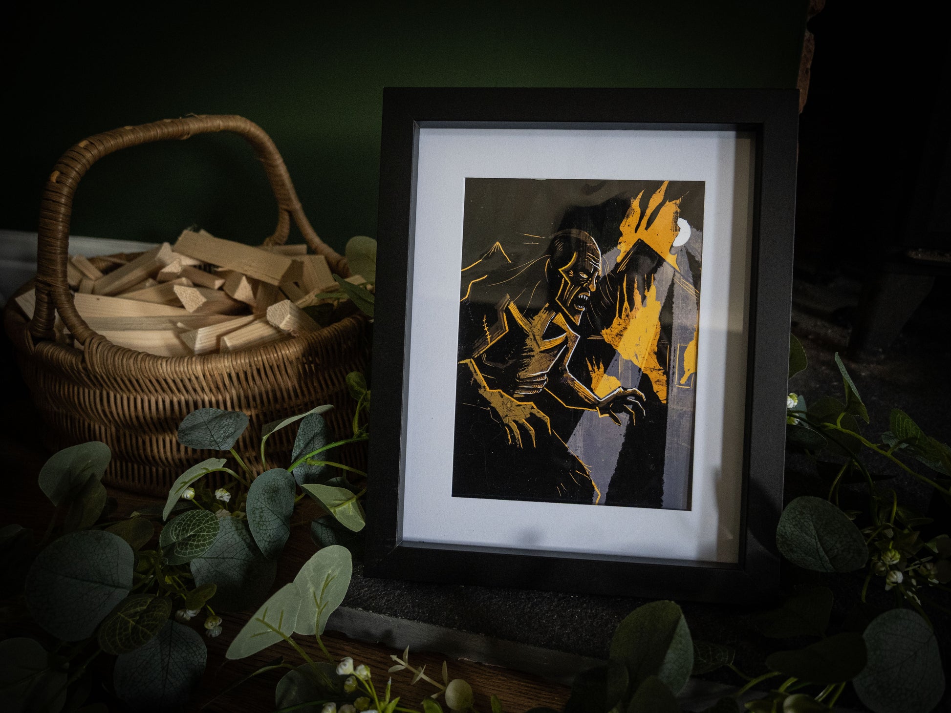 The Creature in the Flames Frankenstein art print in black frame – gaunt monster lunging forward in amber fire, styled with eucalyptus and wicker basket