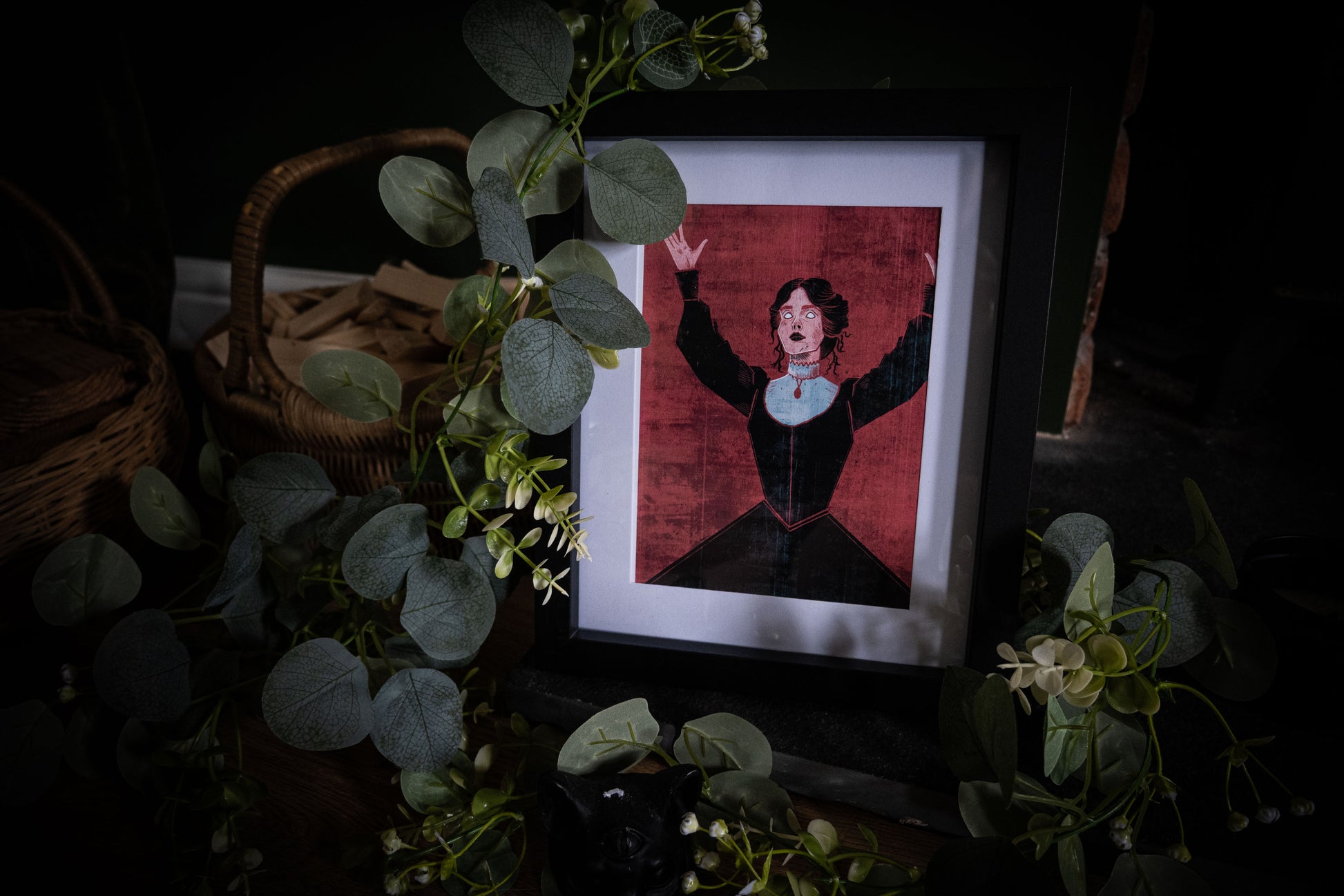 Mina Harker gothic art print by Sophie Atkinson, framed in black on a dark surface surrounded by eucalyptus, arms raised against deep crimson background