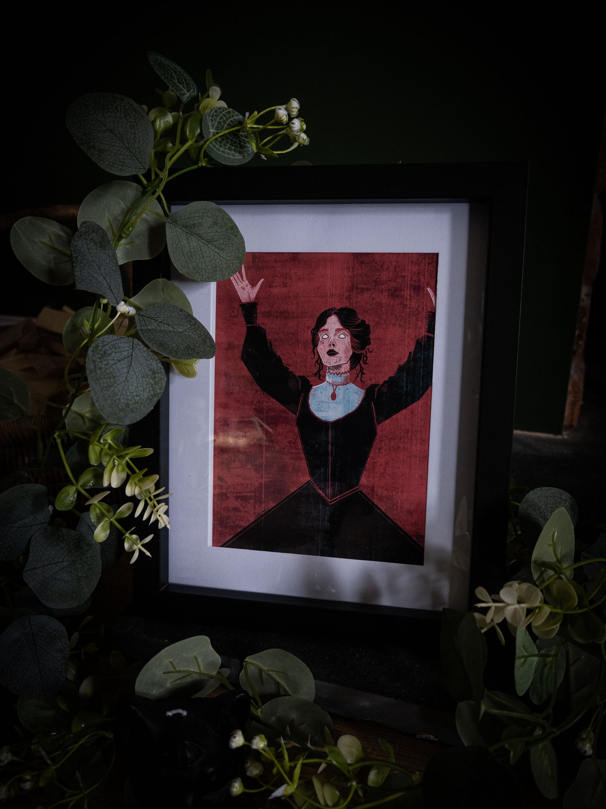 Close-up of Mina Dracula illustration print in black frame, gothic figure in black dress with red choker against crimson wall, styled with botanical foliage