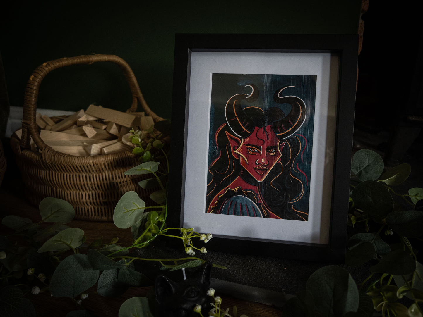 The Horned Temptress gothic demon art print in black frame – red-skinned demoness with curved horns, styled with eucalyptus and wicker basket