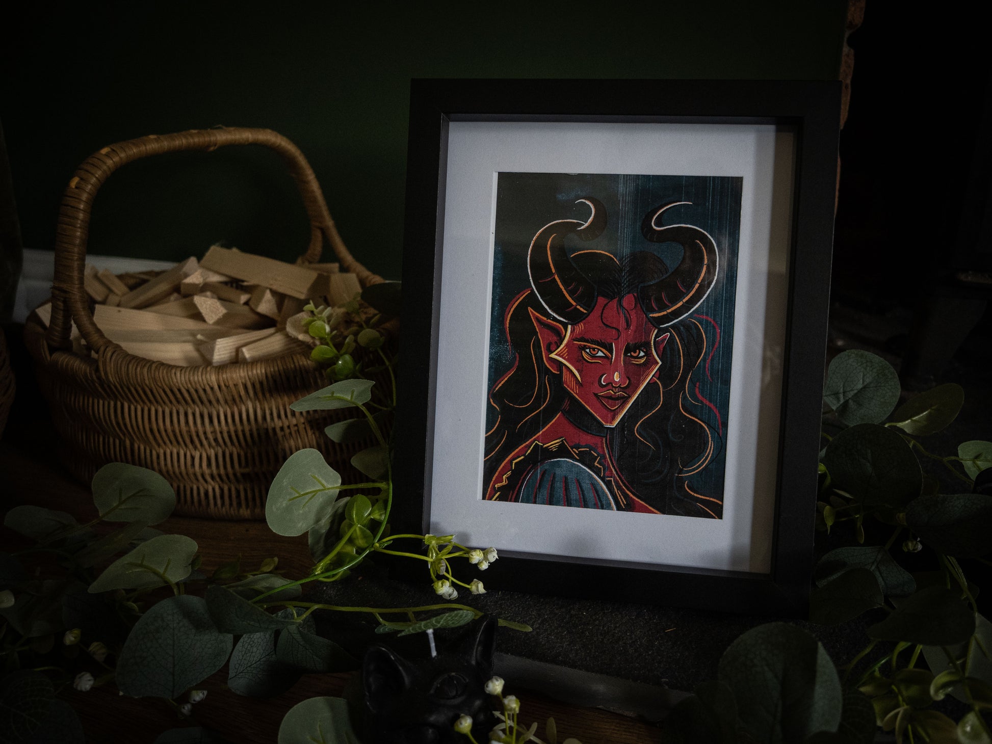 The Horned Temptress gothic demon art print in black frame – red-skinned demoness with curved horns, styled with eucalyptus and wicker basket