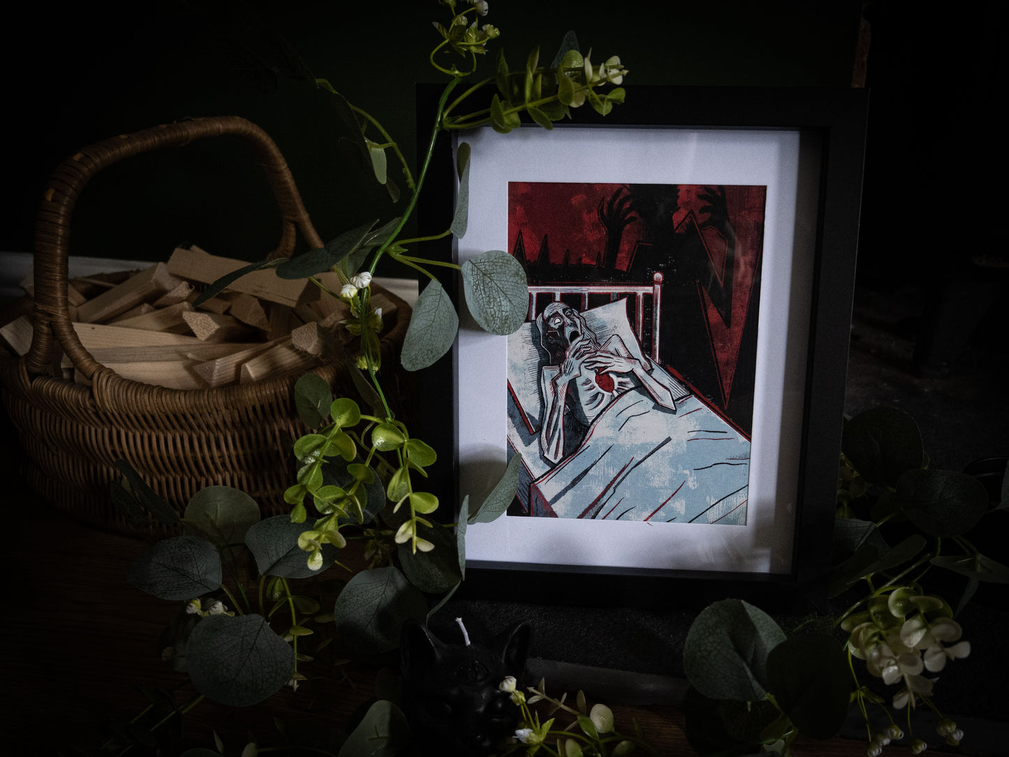 The Tell-Tale Heart gothic art print by Sophie Atkinson, framed in black on a dark wooden surface with wicker basket and eucalyptus, showing gaunt figure clutching a heart with crimson shadow hands looming behind