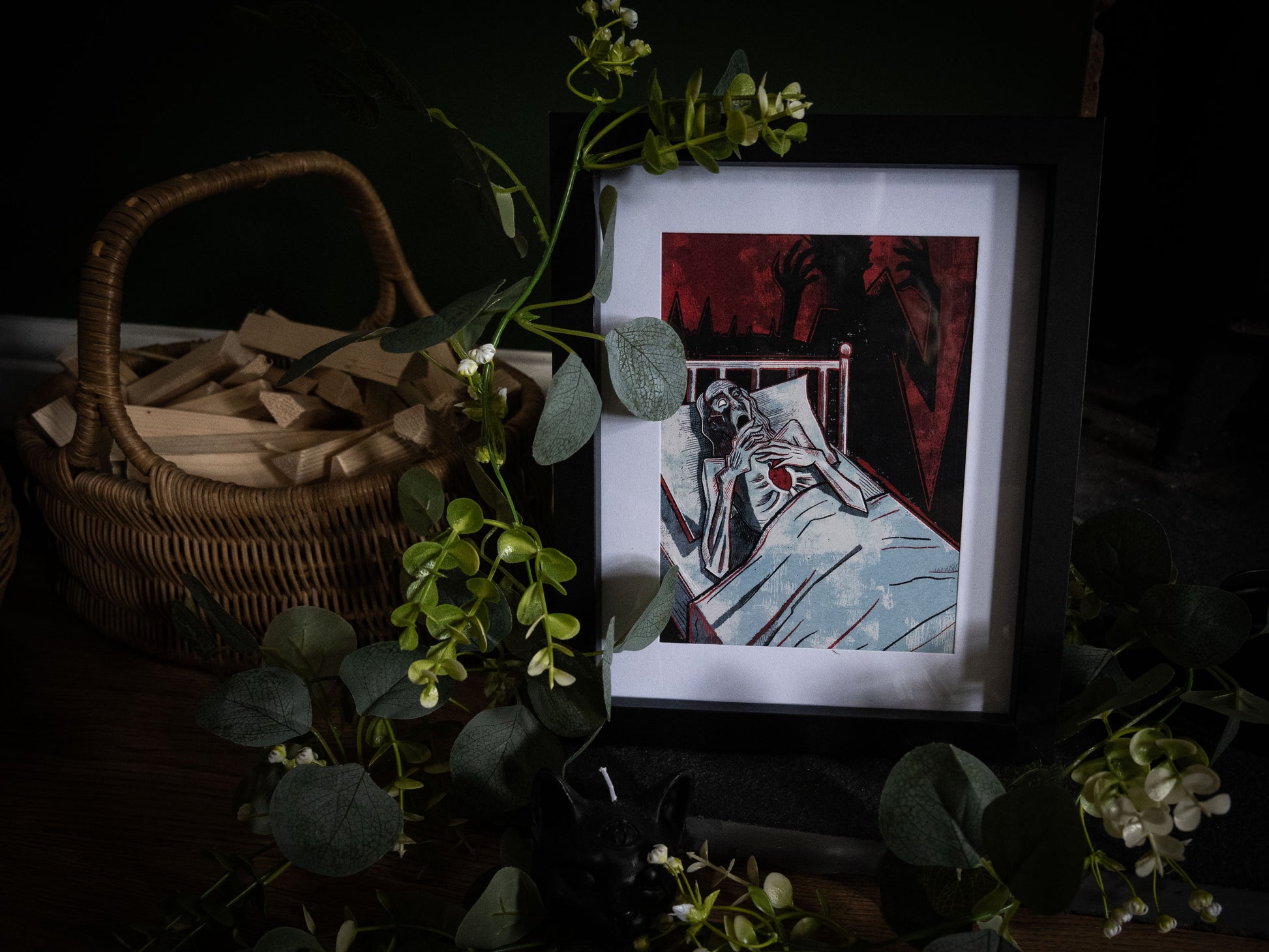 Close-up of The Tell-Tale Heart illustration print in black frame, hollow-eyed figure in bed clutching beating heart, deep crimson and black woodblock-style artwork styled with botanical foliage