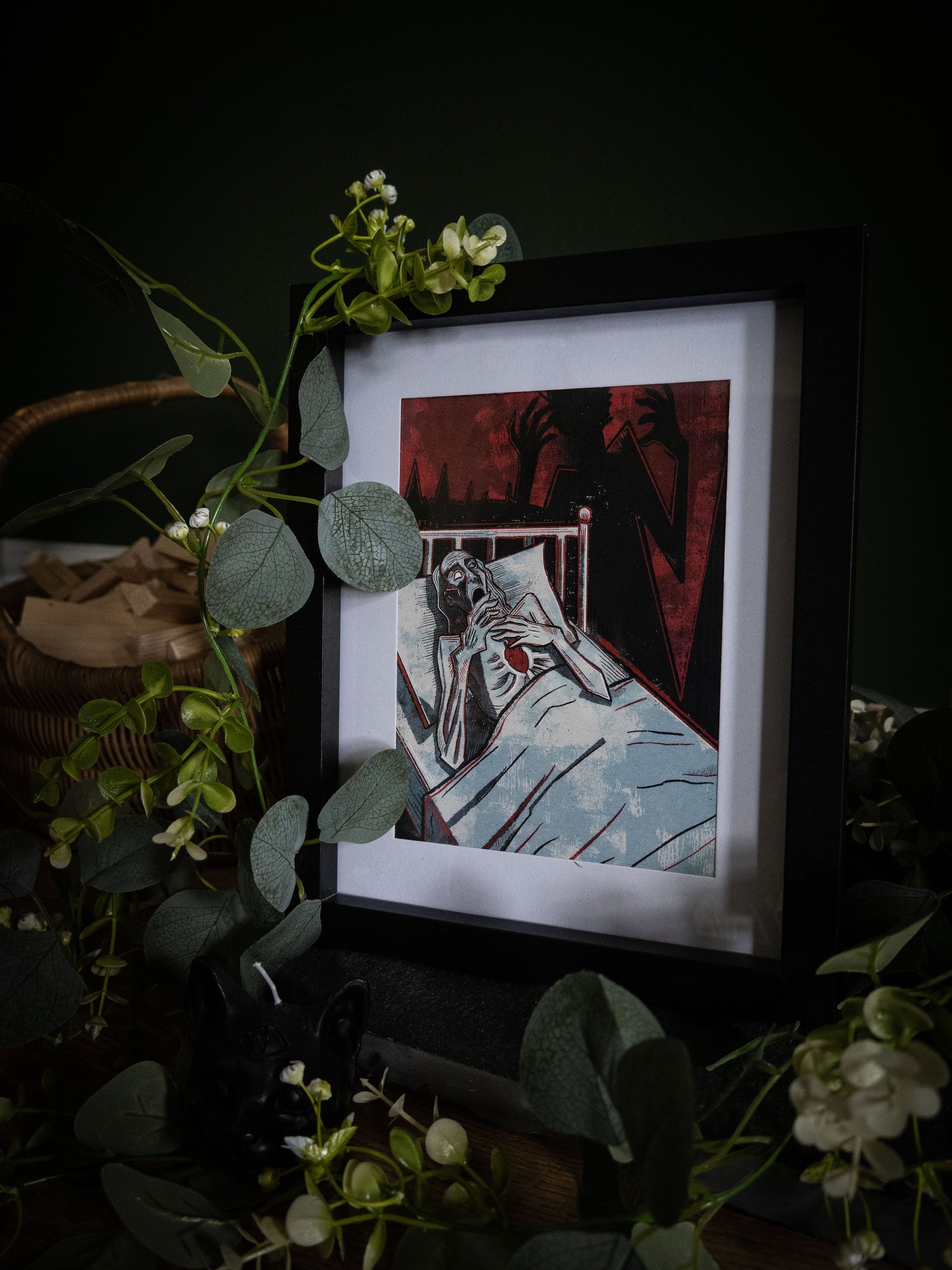 The Tell-Tale Heart Edgar Allan Poe art print in black frame, angled view surrounded by eucalyptus, gaunt figure with crimson shadow hands reaching from dark background