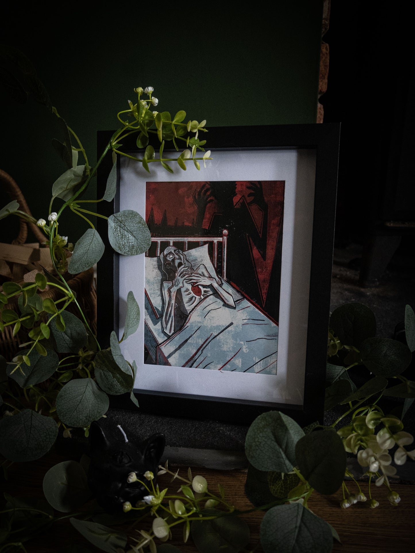 Angled close-up of The Tell-Tale Heart art print in black frame, bold woodblock illustration of figure clutching heart with looming shadow hands, eucalyptus foliage in foreground