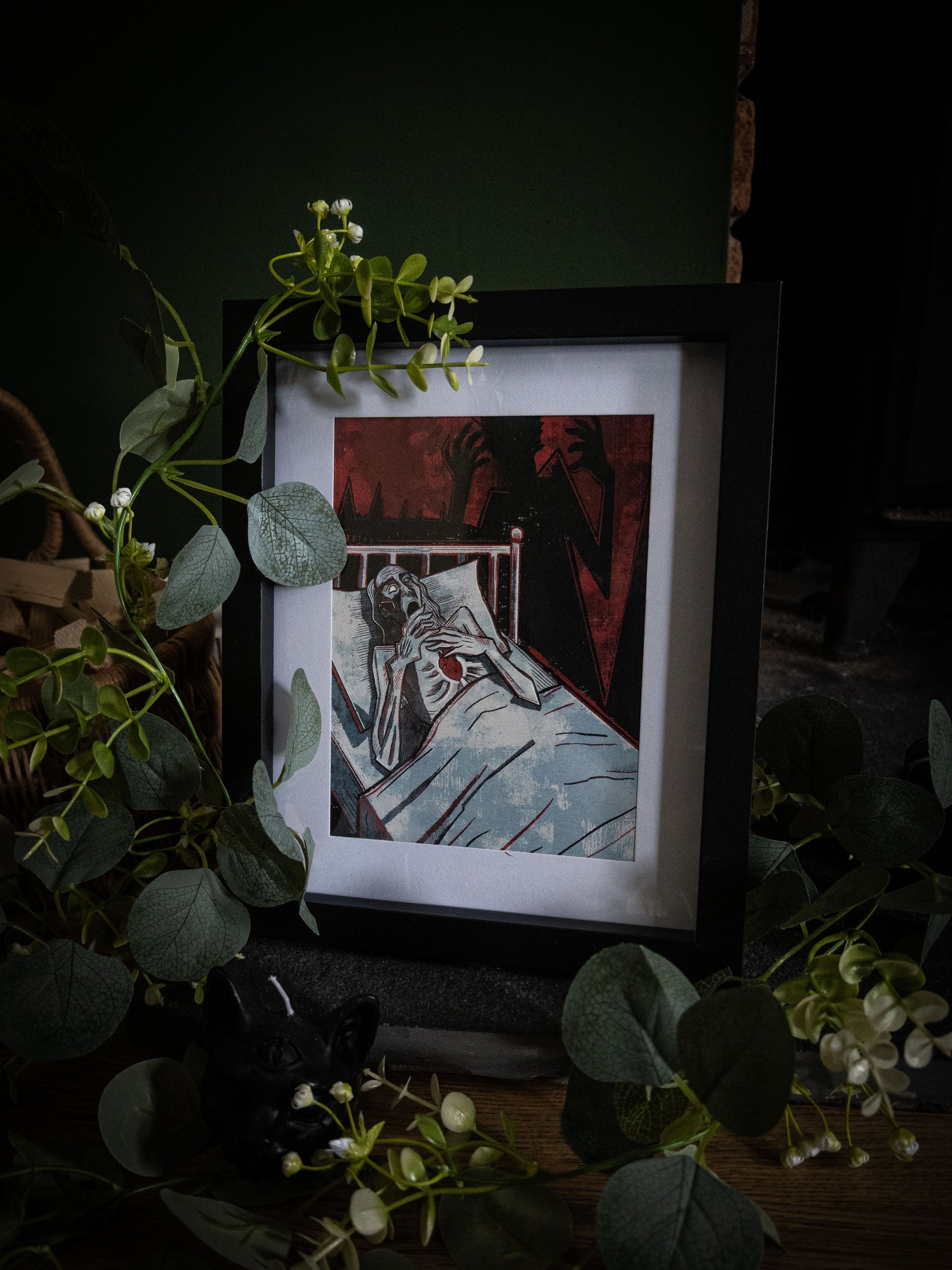 Angled close-up of The Tell-Tale Heart art print in black frame, bold woodblock illustration of figure clutching heart with looming shadow hands, eucalyptus foliage in foreground