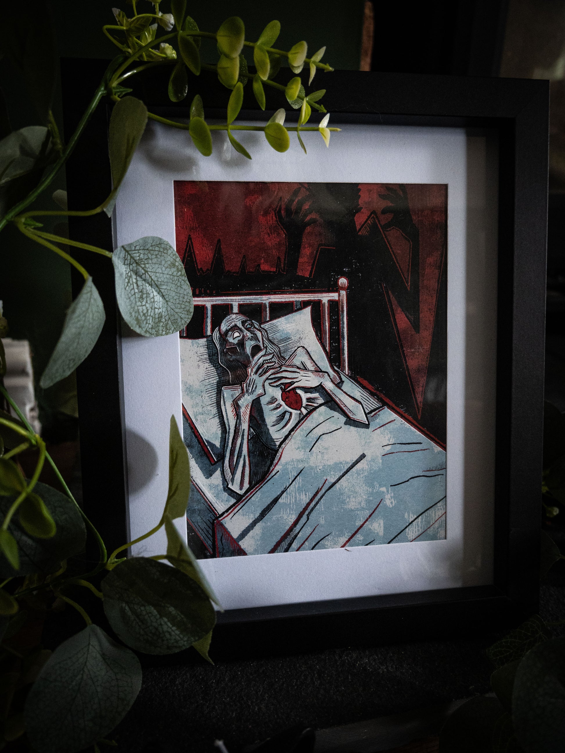 The Tell-Tale Heart gothic fine art print by Sophie Atkinson in black frame, wide styled shot with eucalyptus garland, deep crimson and black illustration visible through white mount