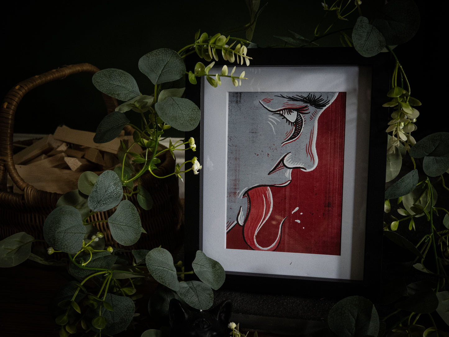 Speak No Evil gothic linocut print – close-up profile face with crimson tongue, framed, surrounded by eucalyptus foliage