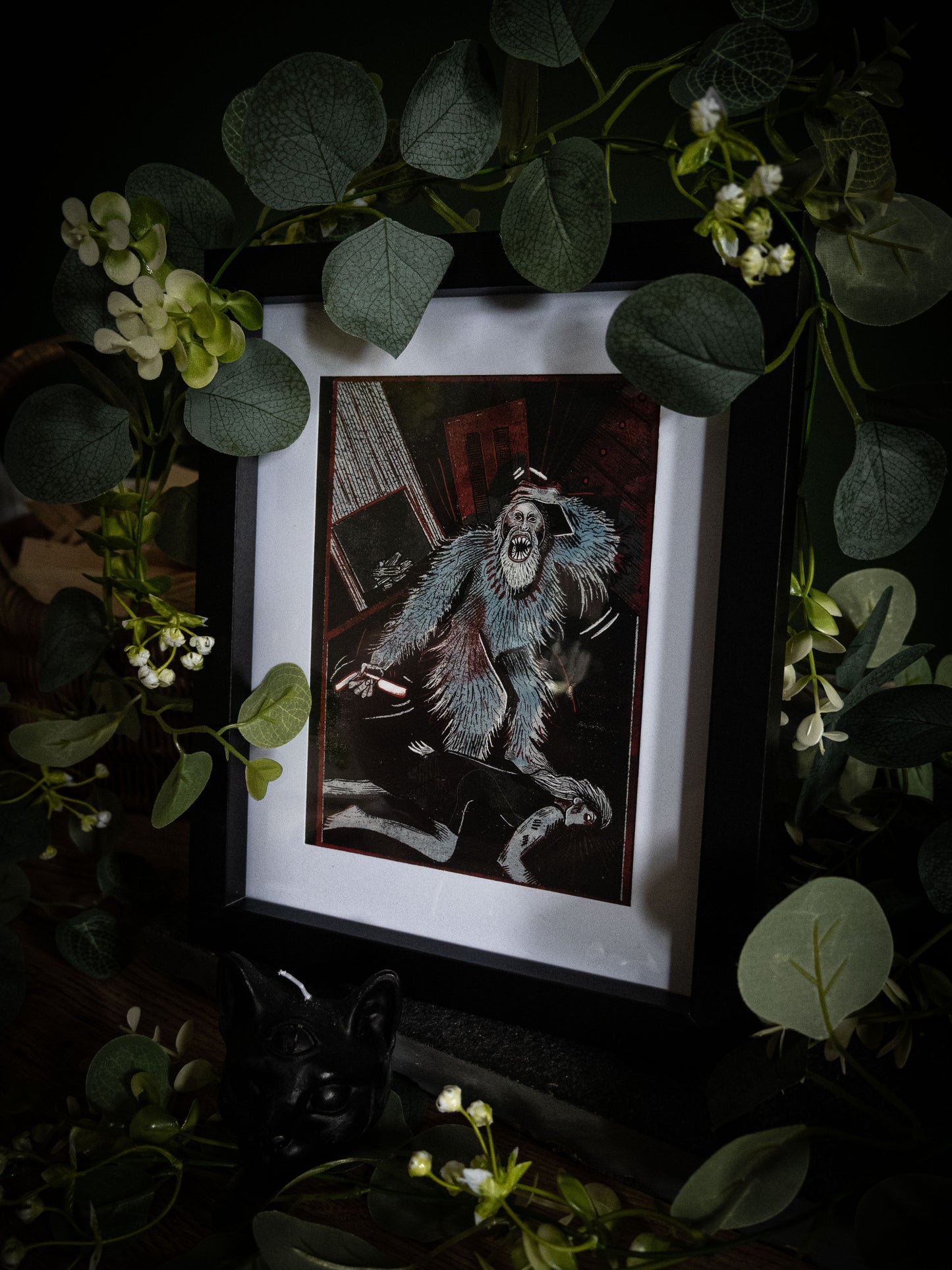 The Murders in the Rue Morgue Edgar Allan Poe art print in black frame, angled view surrounded by eucalyptus garland and white blooms on dark surface