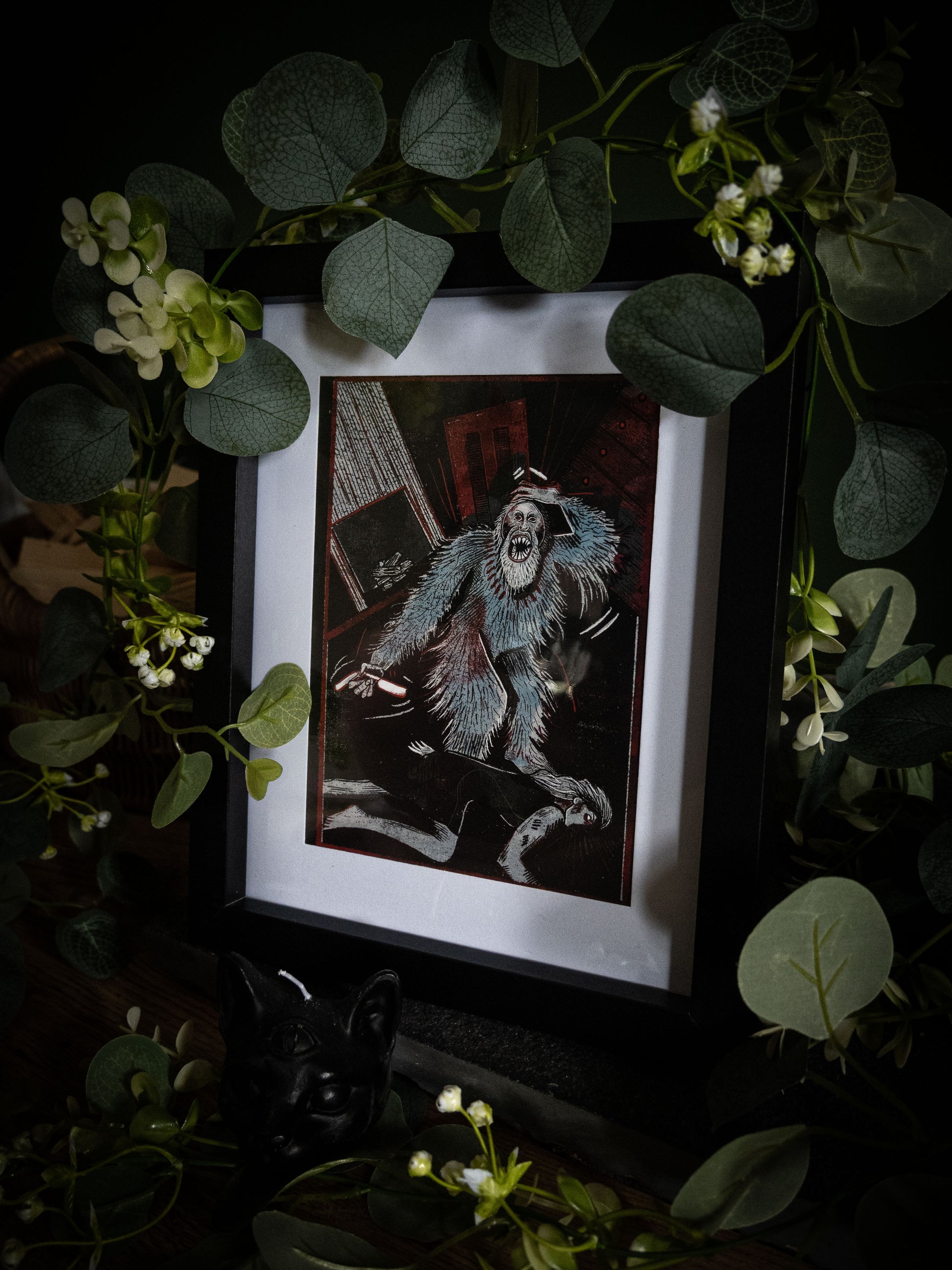 The Murders in the Rue Morgue Edgar Allan Poe art print in black frame, angled view surrounded by eucalyptus garland and white blooms on dark surface